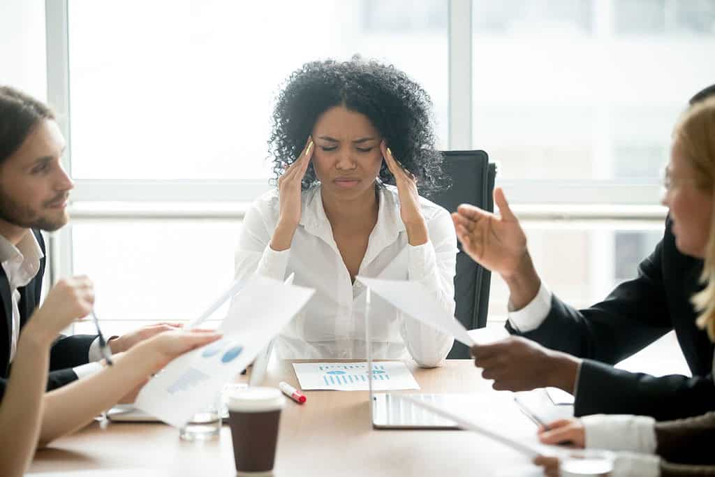 An African American Business Woman With Her Hands on Her Temple During a Meeting with Coworkers Giant Cell Arteritis Treatment